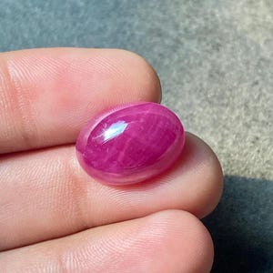 May include: A polished, oval ruby gemstone. The gem is a vibrant, deep pinkish-red colour with a smooth, reflective surface. The stone is held between fingers, showcasing its size and lustre. The gemstone is a cabochon cut.