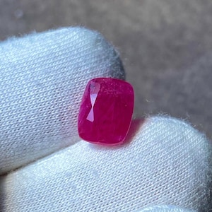 May include: A faceted, cushion-cut ruby gemstone. The ruby is a vibrant, deep red colour, with a polished surface that reflects light. The gemstone is held against a white fabric background.