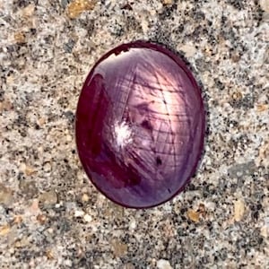 Puede incluir: Una piedra preciosa ovalada de color rojo oscuro con un patrón en forma de estrella en el centro.