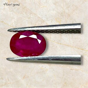May include: An oval-shaped, deep red ruby gemstone held by silver tweezers. The ruby has a faceted top with white reflective surfaces. The background is a light marble pattern, and the text "Hoot gems" is in the upper left corner.