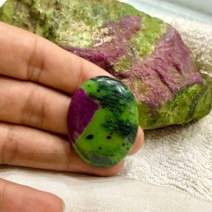 Puede incluir: Gema de zoisita de forma ovalada con una vibrante mezcla de tonos verdes, negros y magenta. La piedra pulida se sostiene cerca de una roca de zoisita rugosa más grande con una coloración similar. El fondo es una superficie blanca texturizada.