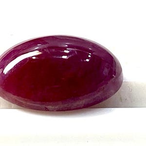 Pode incluir: Uma gema oval de cor vermelha escura em cabochão com uma superfície lisa e polida.