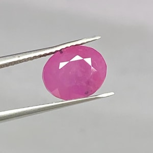 Puede incluir: Una gema rosa de corte ovalado sostenida por unas pinzas de metal plateado. La piedra presenta una superficie facetada que refleja la luz, mostrando un tono rosa intenso y saturado en todo su cuerpo translúcido, con un enfoque claro.