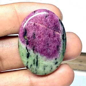 Puede incluir: Gema ovalada con superficie pulida. La piedra presenta una vibrante paleta de colores, con una zona prominente de magenta intenso y una sección verde claro. Salpicaduras negras dispersas.