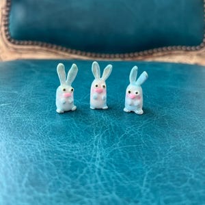 May include: Three small, light blue bunny figurines with pink noses and ears. The bunnies are standing upright, with their ears pointing upwards. The figurines are on a textured, teal surface.