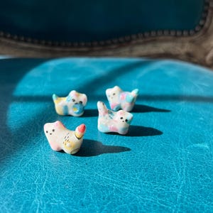 May include: Four small, ceramic cat figurines with pastel color accents. Each cat has a unique design, with pink, blue, yellow, and white hues. The figurines are arranged on a textured, turquoise surface, creating a playful and whimsical scene.