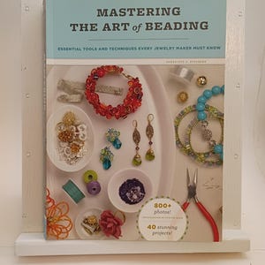 May include: A book titled "Mastering the Art of Beading" displayed in a white wooden stand. The cover features jewelry examples, including bracelets, earrings, and beads. The book promises "Essential Tools and Techniques" with "800+ photos" and "40 stunning projects."