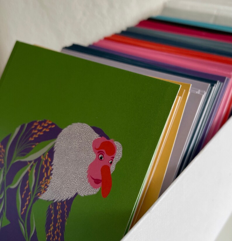 May include: A stack of colorful greeting cards with a green card on top featuring an illustration of a monkey with a pink nose and white fur.