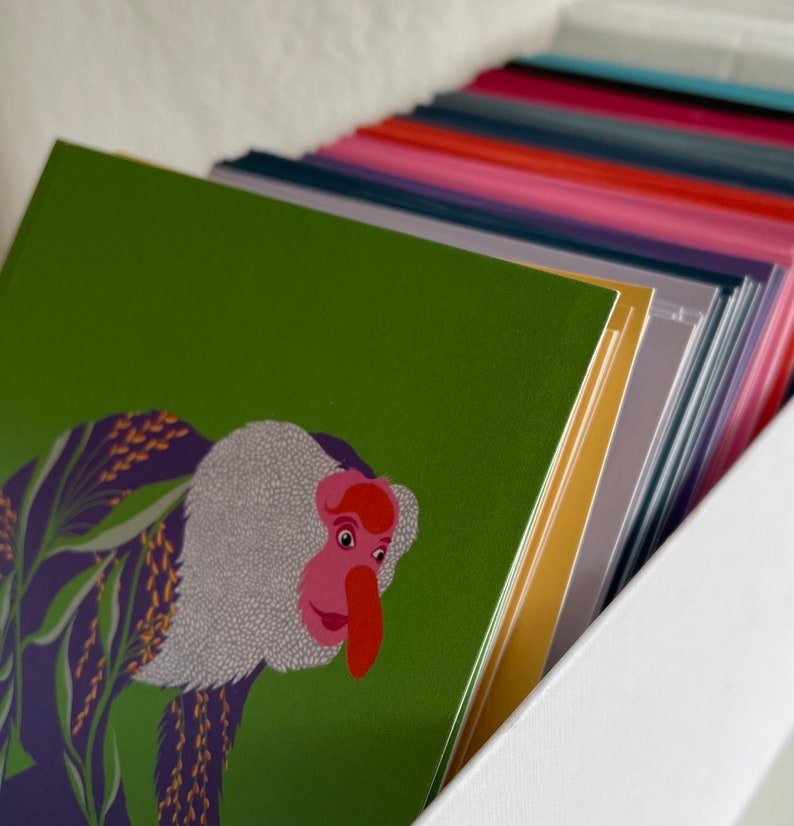 May include: A stack of colorful greeting cards with a green card on top featuring an illustration of a monkey with a pink nose and white fur.
