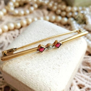 May include: A gold bar pin with two red square-shaped stones and a single pearl in the centre.