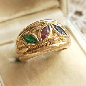 Puede incluir: Un anillo de oro con tres piedras preciosas verdes, rojas y azules, engastadas en un diseño de hojas. El anillo se muestra en una caja blanca con el nombre "ALLARY Pezenas" impreso.