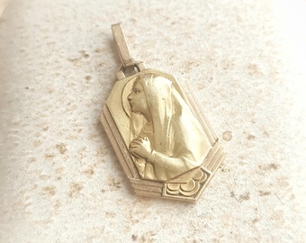 May include: A gold-toned pendant featuring a detailed relief of a woman in profile, set within a geometric frame. The pendant is displayed in a white box.