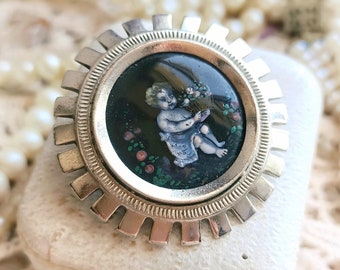 Könnte beinhalten: Eine silberfarbene Brosche mit einem runden, schwarzen Emaille-Kamee, der einen Cherub mit einem Blumenstrauß zeigt. Der Kamee ist von einem dekorativen, gewellten Rand umgeben.