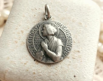 May include: Silver pendant featuring a religious image of a woman with a halo, surrounded by a decorative border with the text "SANCTA SOLENGISTO".