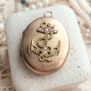 Puede incluir: Medallón de oro con un diseño de ancla, adornado con piedras preciosas rojas y blancas. El medallón se muestra sobre una superficie blanca.