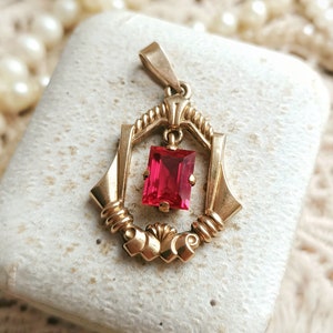 May include: A gold pendant with a red gemstone in a geometric setting. The pendant is in a small white box.