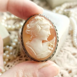 Peut inclure: Une broche camée représentant un portrait sculpté d'une femme en robe blanche avec un motif floral. Le camée est monté dans un cadre ovale argenté avec un bord perlé.