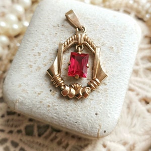 May include: A gold pendant with a red gemstone set in a geometric design. The pendant is in a small white box.