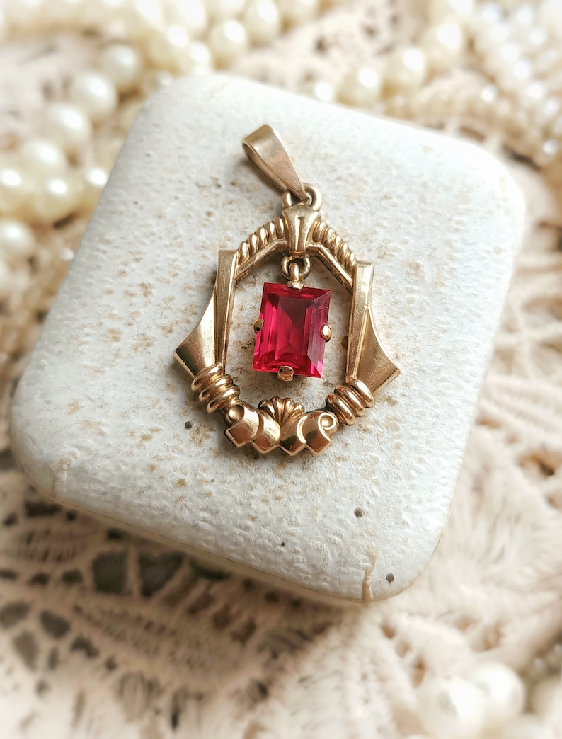 May include: A gold pendant with a red gemstone set in a geometric design. The pendant is in a small white box.
