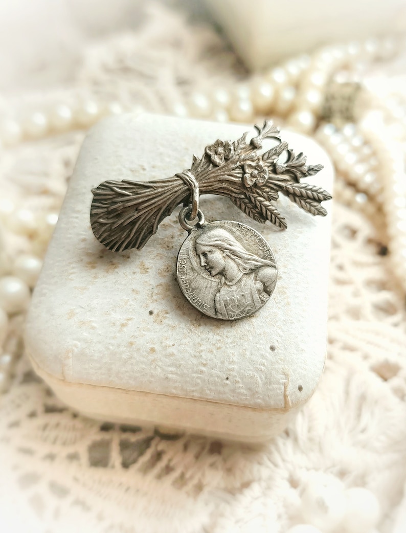 May include: A silver charm featuring a bouquet of wheat and a round medal with a depiction of the Virgin Mary. The medal has the text "Ave Maria" around the edge.