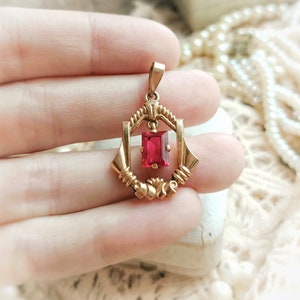 May include: A gold pendant with a red gemstone in a geometric setting. The pendant is held in a hand.