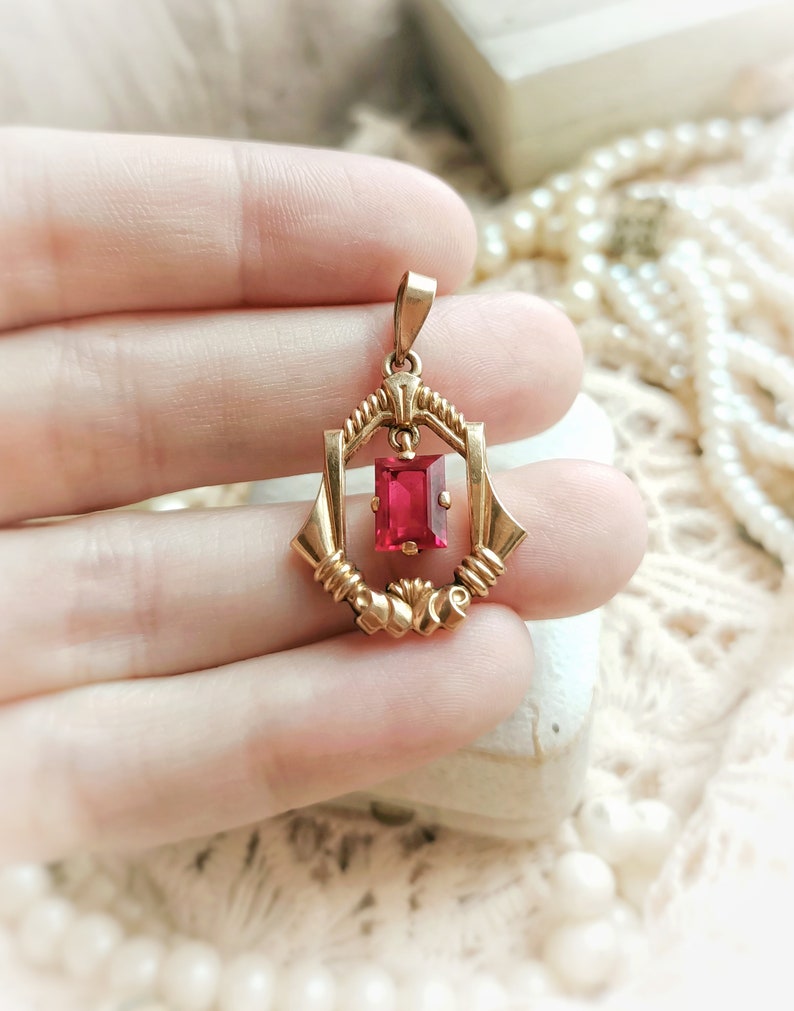 May include: A gold pendant with a red gemstone in a geometric setting. The pendant is held in a hand.