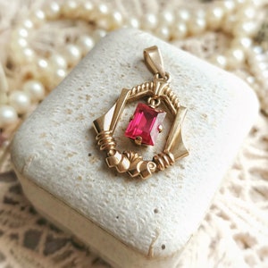 May include: A gold pendant with a red gemstone in a geometric design. The pendant is in a white box.