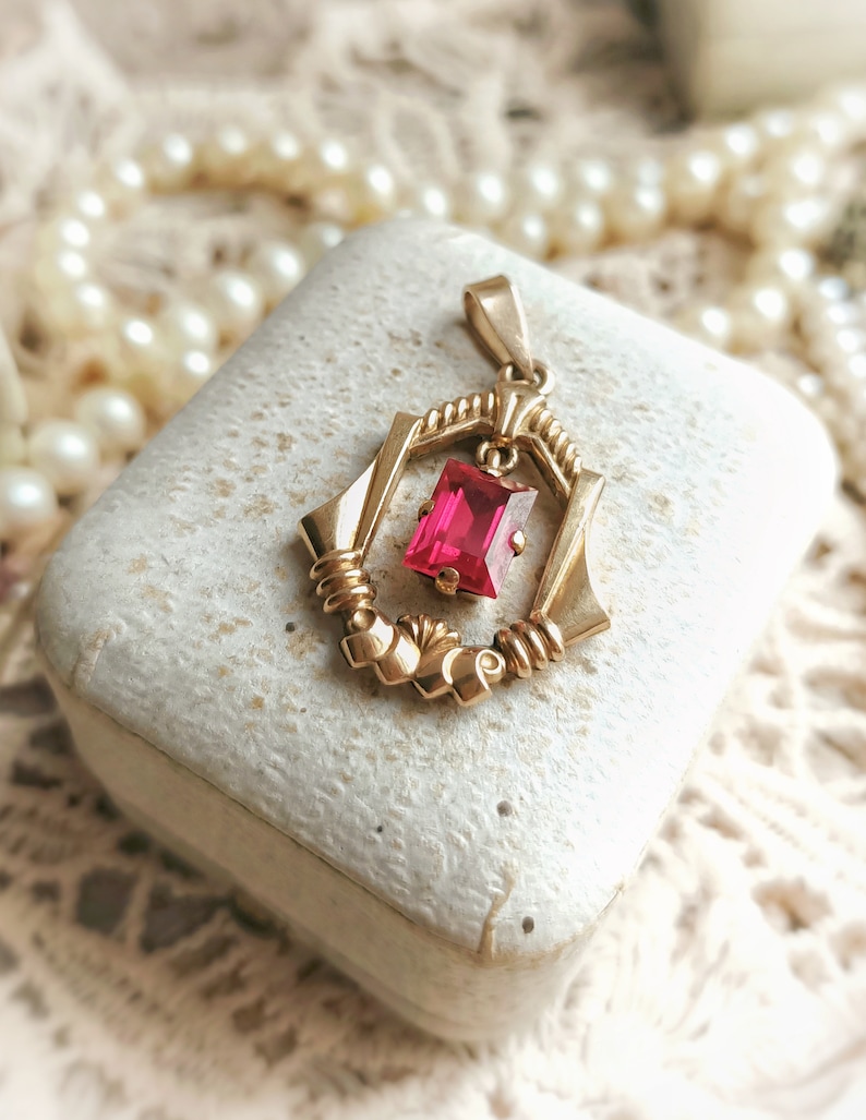 May include: A gold pendant with a red gemstone in a geometric design. The pendant is in a white box.