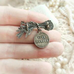 May include: A silver-toned brooch with a floral design and a round pendant that reads "VIVE LABEUR".