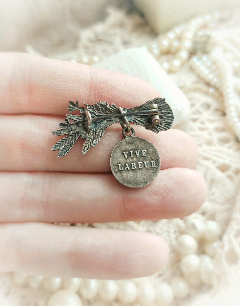 May include: A silver-toned brooch with a floral design and a round pendant that reads "VIVE LABEUR".