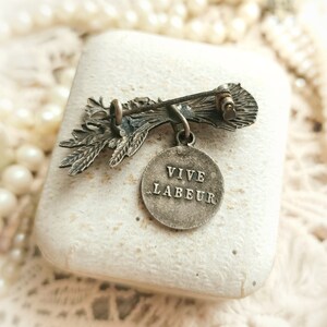 May include: Silver tone brooch featuring a stylised floral design with a round pendant that reads "VIVE LABEUR".