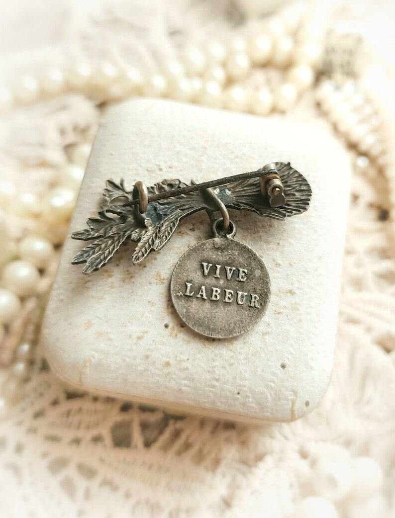 May include: Silver tone brooch featuring a stylised floral design with a round pendant that reads "VIVE LABEUR".