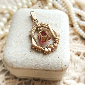 May include: A gold pendant with a red gemstone hanging from a delicate, ornate frame. The pendant is in a white box.