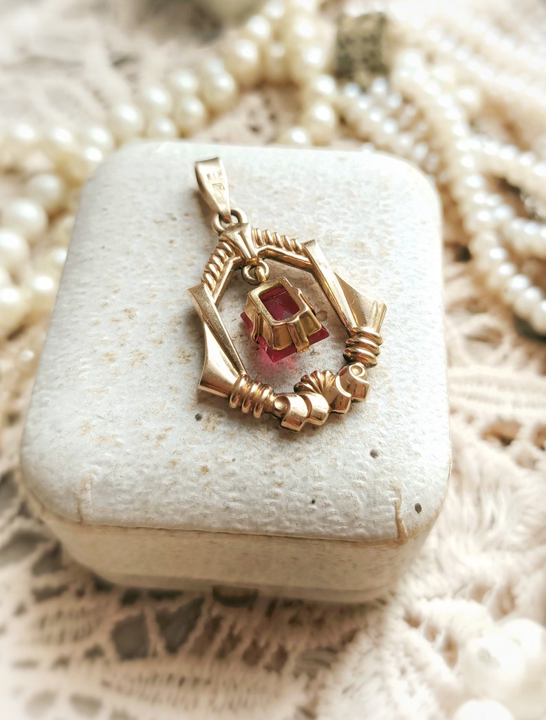 May include: A gold pendant with a red gemstone hanging from a delicate, ornate frame. The pendant is in a white box.