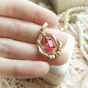 May include: A gold pendant with a pink gemstone set in a geometric frame. The pendant is held in a hand.