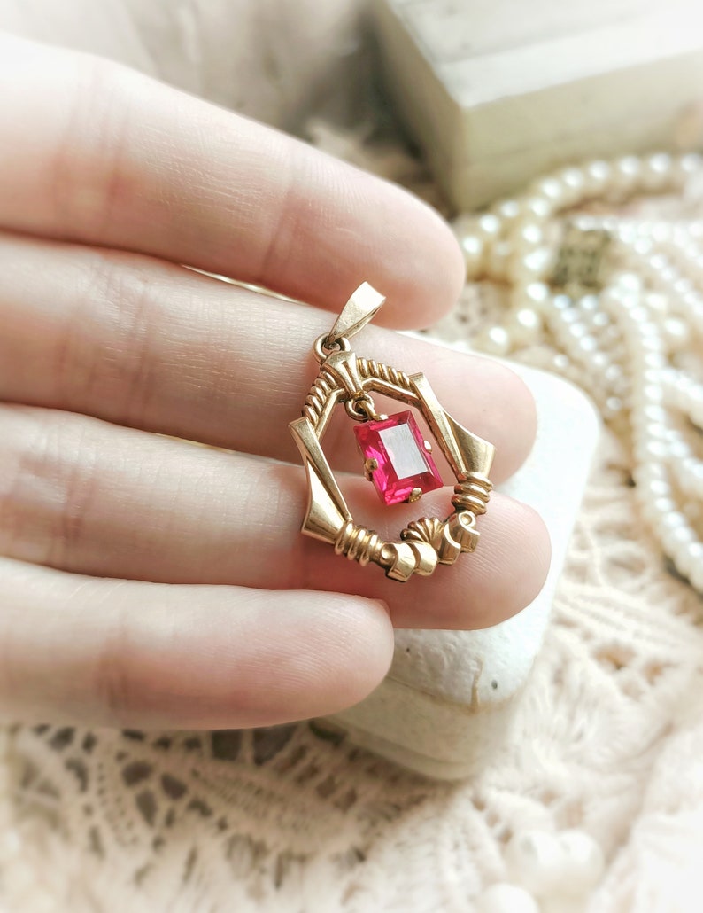 May include: A gold pendant with a pink gemstone set in a geometric frame. The pendant is held in a hand.