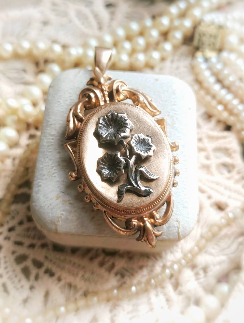 K&ouml;nnte beinhalten: Ein goldenes Medaillon mit einem floralen Design. Das Medaillon ist oval geformt und hat ein detailliertes florales Design mit drei Blumen und Bl&auml;ttern. Die Blumen sind silber und die Bl&auml;tter sind golden.