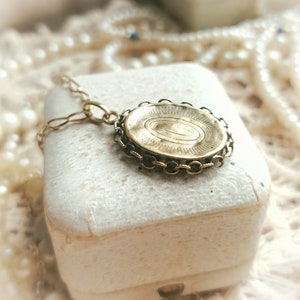 May include: A gold locket necklace with a detailed engraved design on the front. The locket is in a white box and surrounded by pearls.
