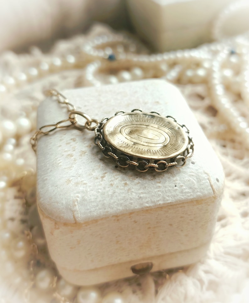 May include: A gold locket necklace with a detailed engraved design on the front. The locket is in a white box and surrounded by pearls.