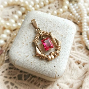May include: A gold pendant with a red gemstone in a geometric design. The pendant is in a small white box.