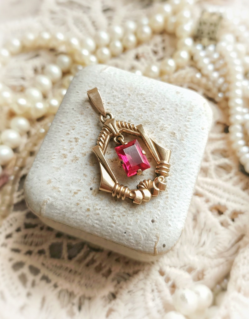 May include: A gold pendant with a red gemstone in a geometric design. The pendant is in a small white box.