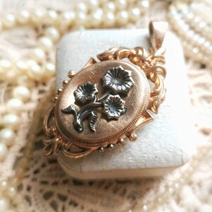 K&ouml;nnte beinhalten: Ein goldenes Medaillon mit einem floralen Design. Das Medaillon ist oval geformt und hat auf der Vorderseite ein zartes florales Design. Die Blumen sind aus Silber und auf einem goldenen Hintergrund angebracht.