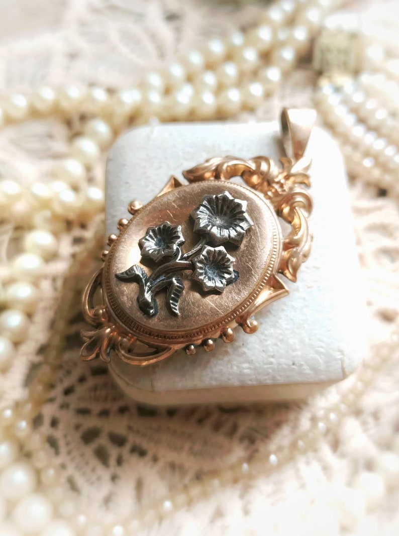 K&ouml;nnte beinhalten: Ein goldenes Medaillon mit einem floralen Design. Das Medaillon ist oval geformt und hat auf der Vorderseite ein zartes florales Design. Die Blumen sind aus Silber und auf einem goldenen Hintergrund angebracht.