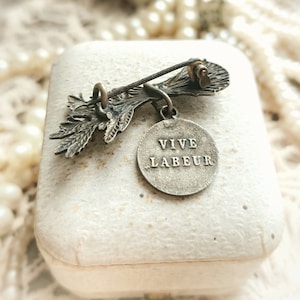 May include: A silver-toned brooch with a floral design and a round pendant that reads "VIVE LABEUR".