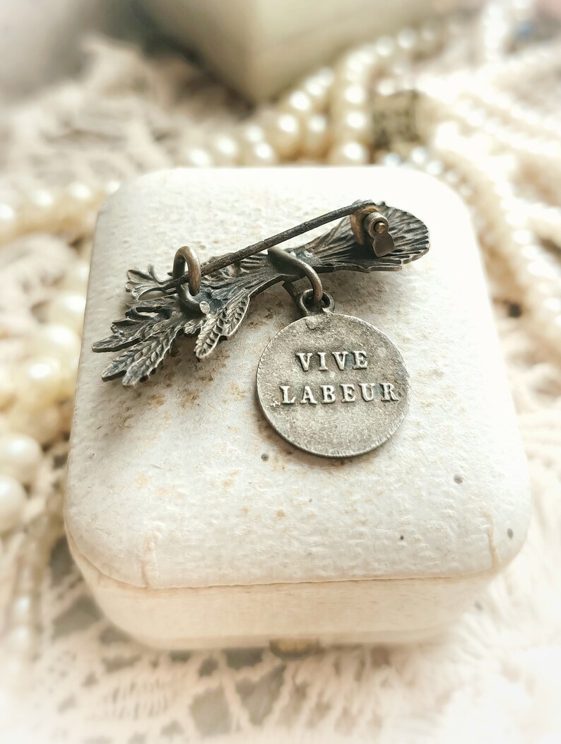 May include: A silver-toned brooch with a floral design and a round pendant that reads "VIVE LABEUR".