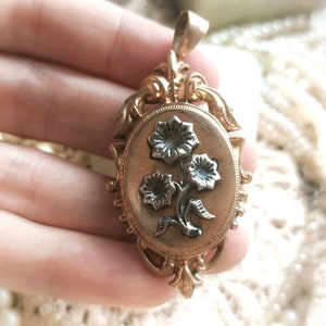 K&ouml;nnte beinhalten: Ein goldenes Medaillon mit einem verzierten Blumenmuster. Das Medaillon zeigt drei Blumen mit schwarzen Zentren und goldenen Bl&uuml;tenbl&auml;ttern.