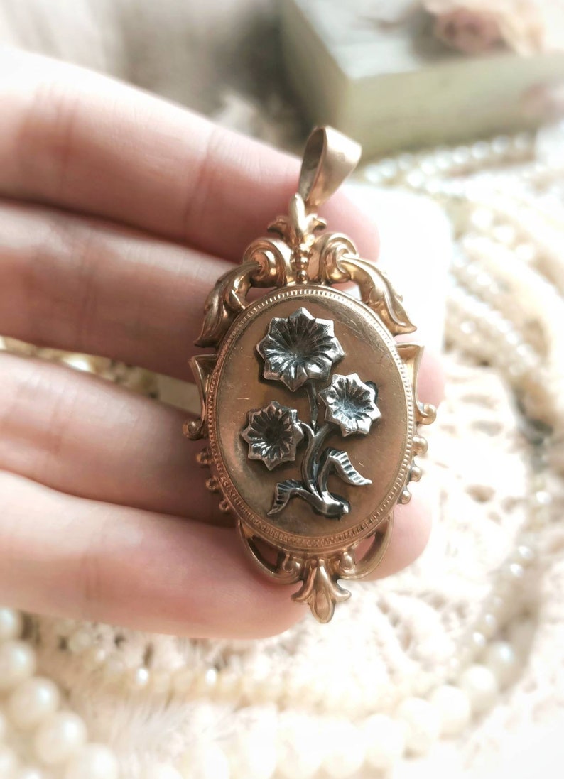 K&ouml;nnte beinhalten: Ein goldenes Medaillon mit einem verzierten Blumenmuster. Das Medaillon zeigt drei Blumen mit schwarzen Zentren und goldenen Bl&uuml;tenbl&auml;ttern.