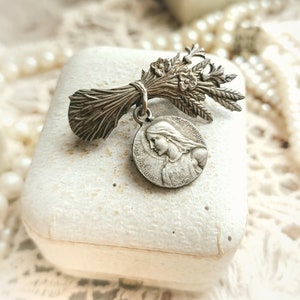 May include: Silver tone religious brooch featuring a bouquet of wheat and a round medal with an image of the Virgin Mary. The medal has the text "JESUS MARIA" around the edge.