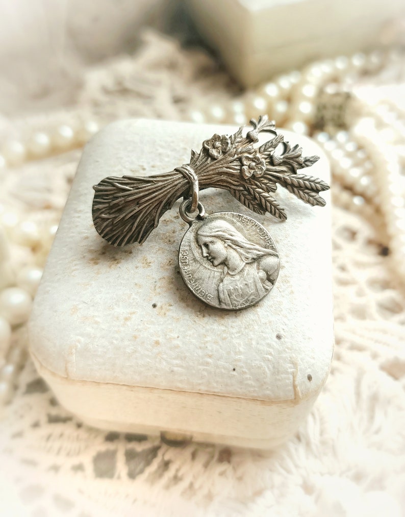 May include: Silver tone religious brooch featuring a bouquet of wheat and a round medal with an image of the Virgin Mary. The medal has the text "JESUS MARIA" around the edge.