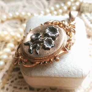 K&ouml;nnte beinhalten: Ein goldenes Medaillon mit einem floralen Design. Das Medaillon ist oval geformt und hat auf der Vorderseite ein zartes Blumenmuster. Die Blumen sind aus Silber und haben einen schwarzen Emaille-Mittelpunkt.